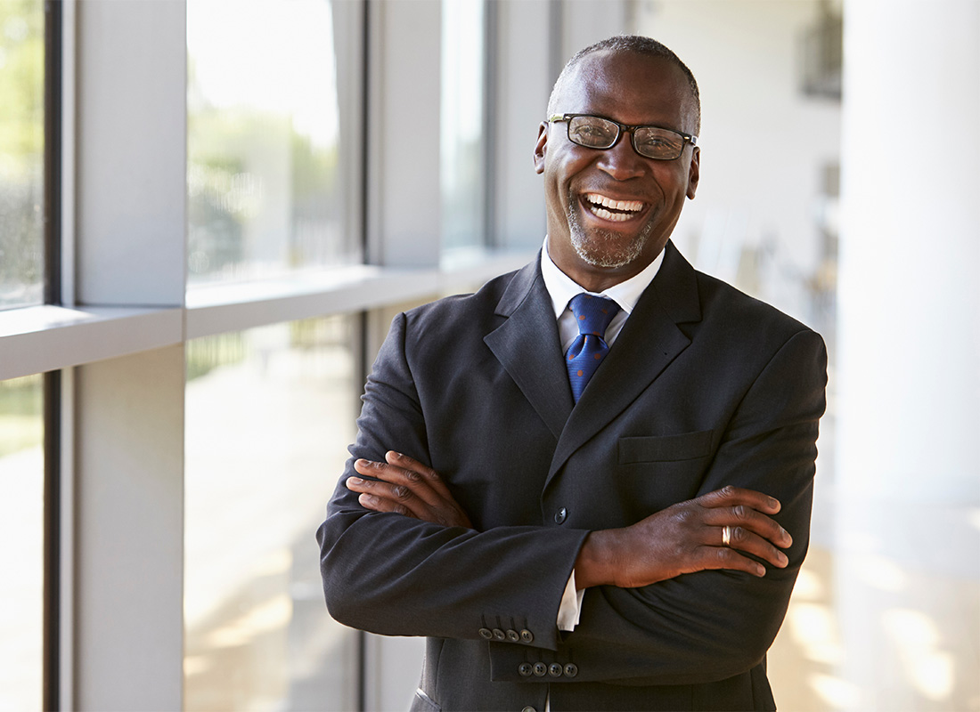 Business Insurance - Portrait of a Middle Aged Smiling Businessman Wearing Glasses Standing Next to Bright Windows in a Modern Office Building with his Arms Folded