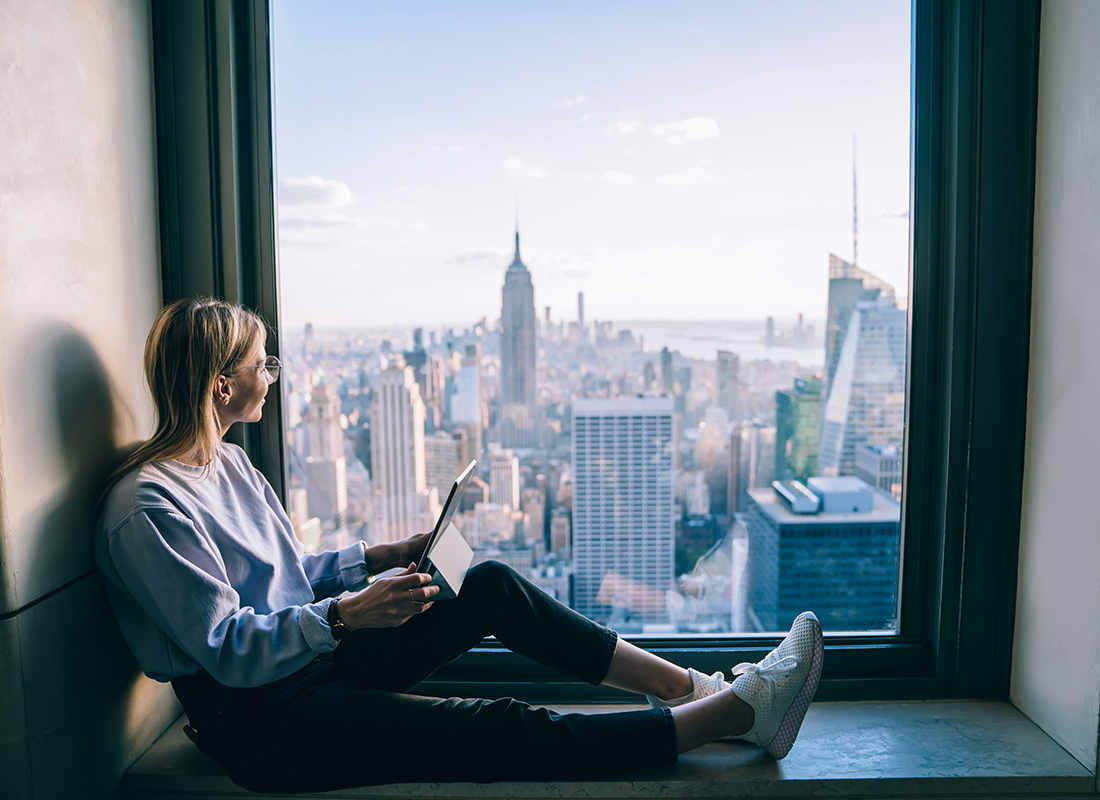 Contact - View of a Young Woman Holding a Tablet Sitting Next to a Large Window in a Tall Building with Views of Downtown Manhattan